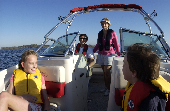 Mom and dad, wearing inflatable life jackets, with kids in the bow with the shoreline in the background.