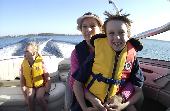 Male child sitting on adult female's lap with young girl in a children's life jacket sitting in the back. The shoreline is in the background.