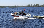 Adult male (dad) and adult female (mom) on motor boat with children (boy and girl) sitting in the bow. The shoreline is in the background.