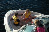 Boy and girl wearing children's life jackets while in the bow, while an adult female wearing an inflatable life jackets watches over them