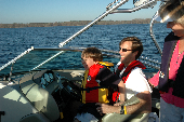 Adult male wearing an inflatable life jacket, with a boy wearing a children's life jacket sitting on his lap while an female looks on. The shoreline is in the background.