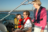 Adult male wearing an inflatable life jacket, with a boy wearing a children's life jacket sitting on his lap while a female looks on. The shoreline is in the background.