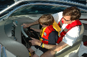 Man wearing inflatable life jacket and boy wearing children's life jacket sitting behind the steering wheel of a motor boat.